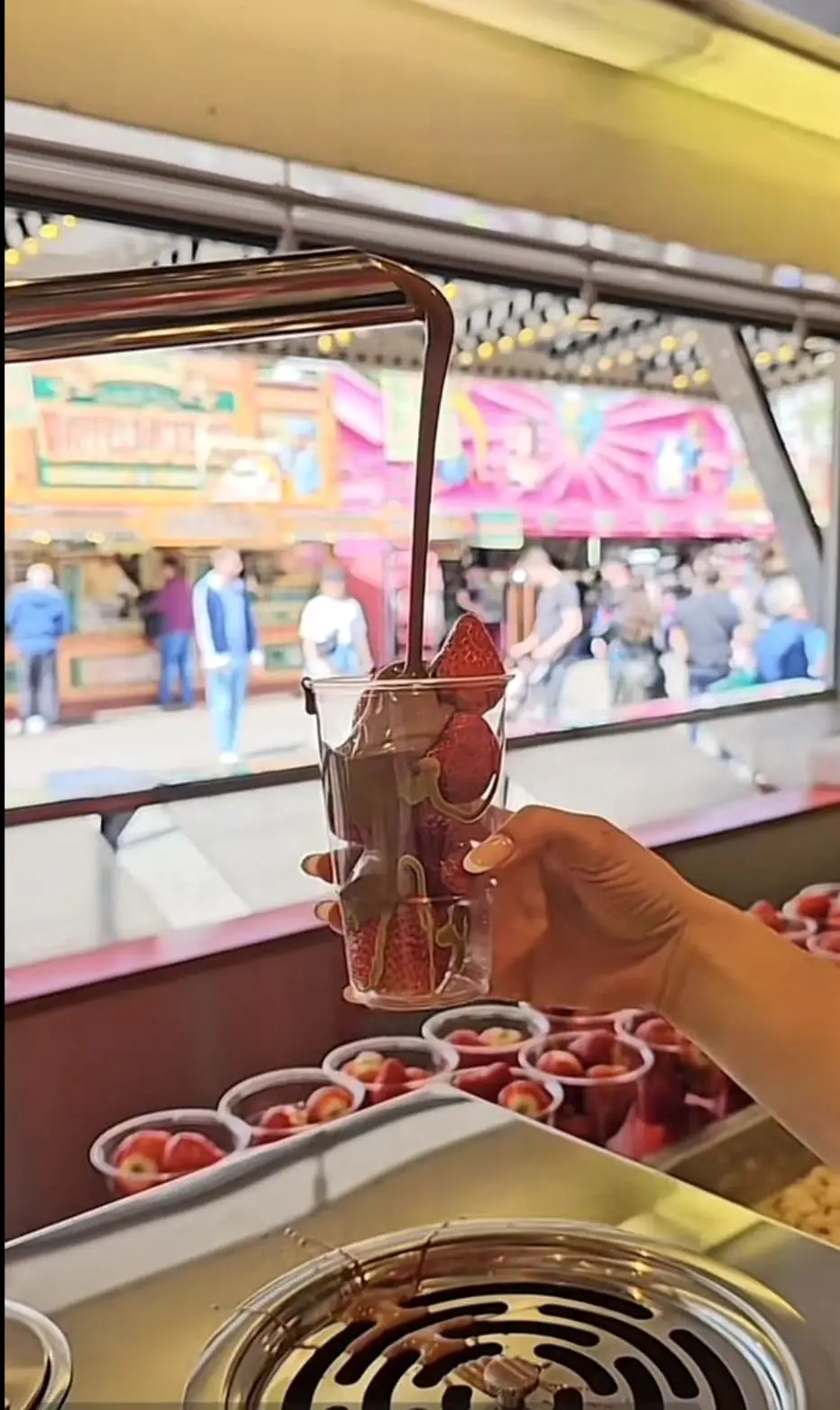 Ein Becher mit Erdbeeren und Schokolade in der Hand, dahinter das Festgelände