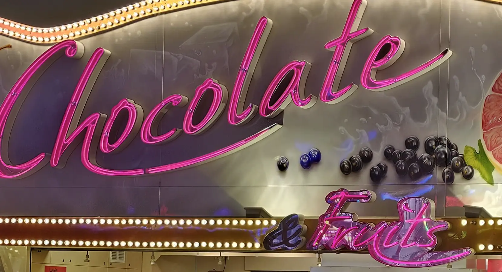Nahaufnahme des Neonschriftzugs Chocolate & Fruits mit Glühbirnenkette darunter