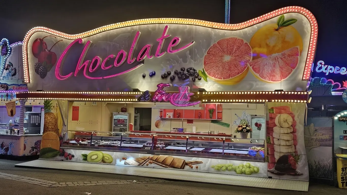 Der Wagen Chocolate & Fruits am Abend auf dem Festplatz, der pinke Neonschriftzug leuchtet über der Theke